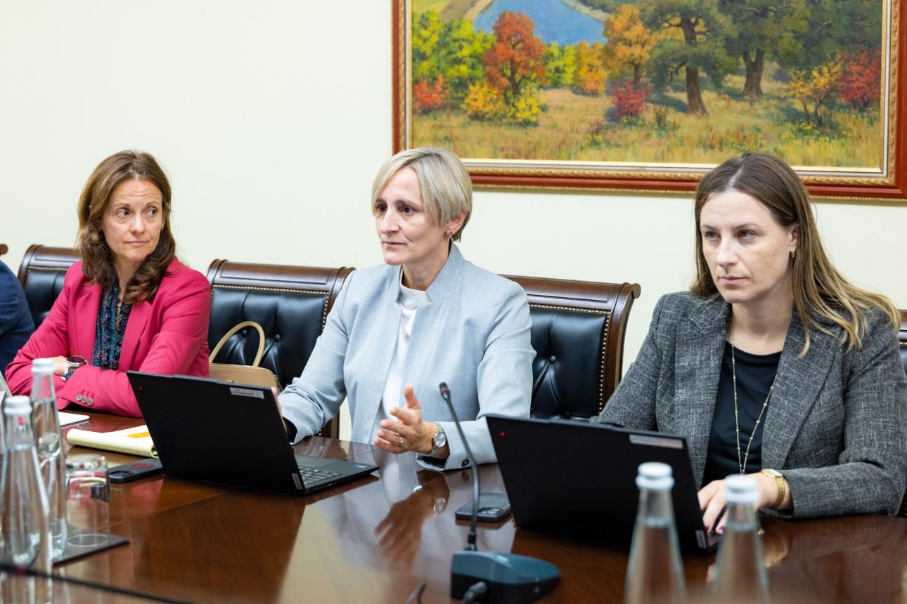 Trei femei așezate la o masă de ședințe, cu laptopuri deschise, una dintre ele gesticulând în timp ce vorbește
