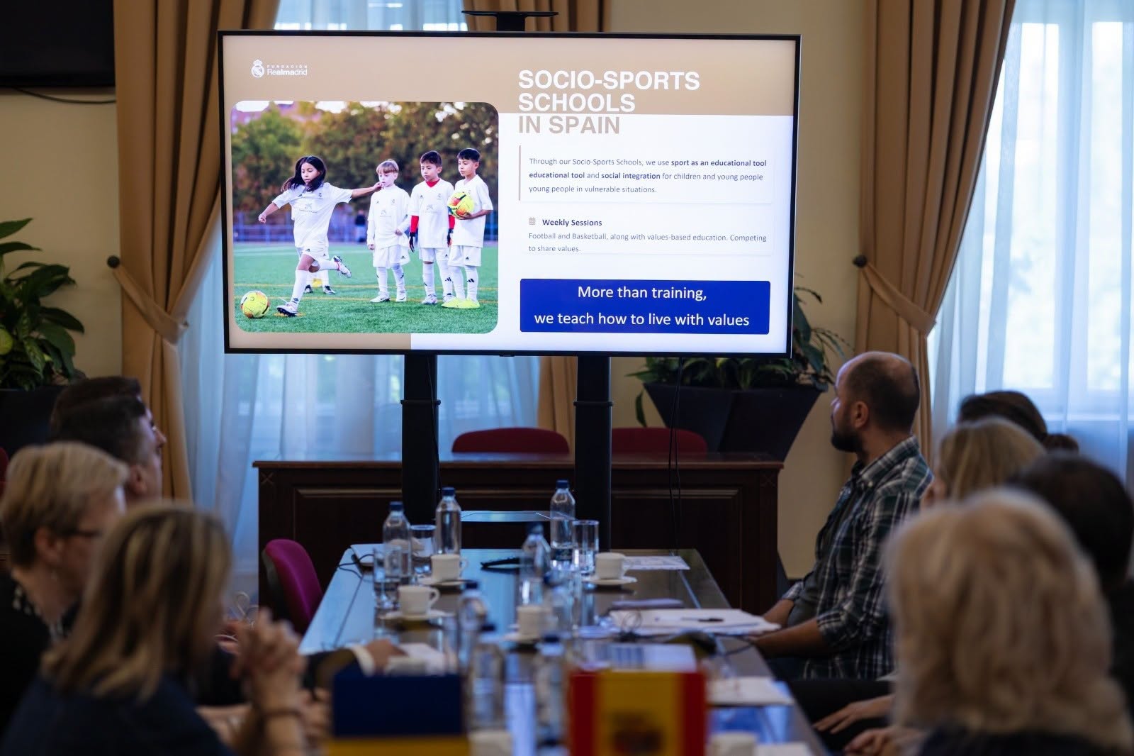 Participanți la o masă urmăresc pe un ecran prezentarea școlilor sociale sportive ale Fundației Real Madrid