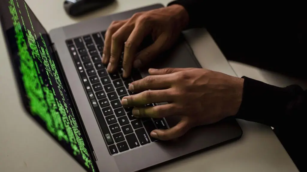 Mâini care tastează pe tastatura unui laptop, cu rânduri de cod verde pe ecran
